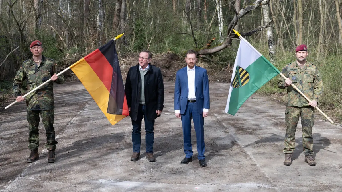 27.03.2024, Sachsen, Bernsdorf: Boris Pistorius (SPD, l), Bundesverteidigungsminister, und Michael Kretschmer (CDU), Ministerpräsident von Sachsen, stehen auf dem Gelände eines geplanten Bundeswehrstandorts zwischen einer Deutschlandfahne und einer Fahne des Freistaat Sachsen. Auf dem Areal in Bernsdorf-Straßgräbchen soll künftig das neu aufzustellende Logistikbataillon 471 stationiert werden. Foto: Sebastian Kahnert/dpa +++ dpa-Bildfunk +++