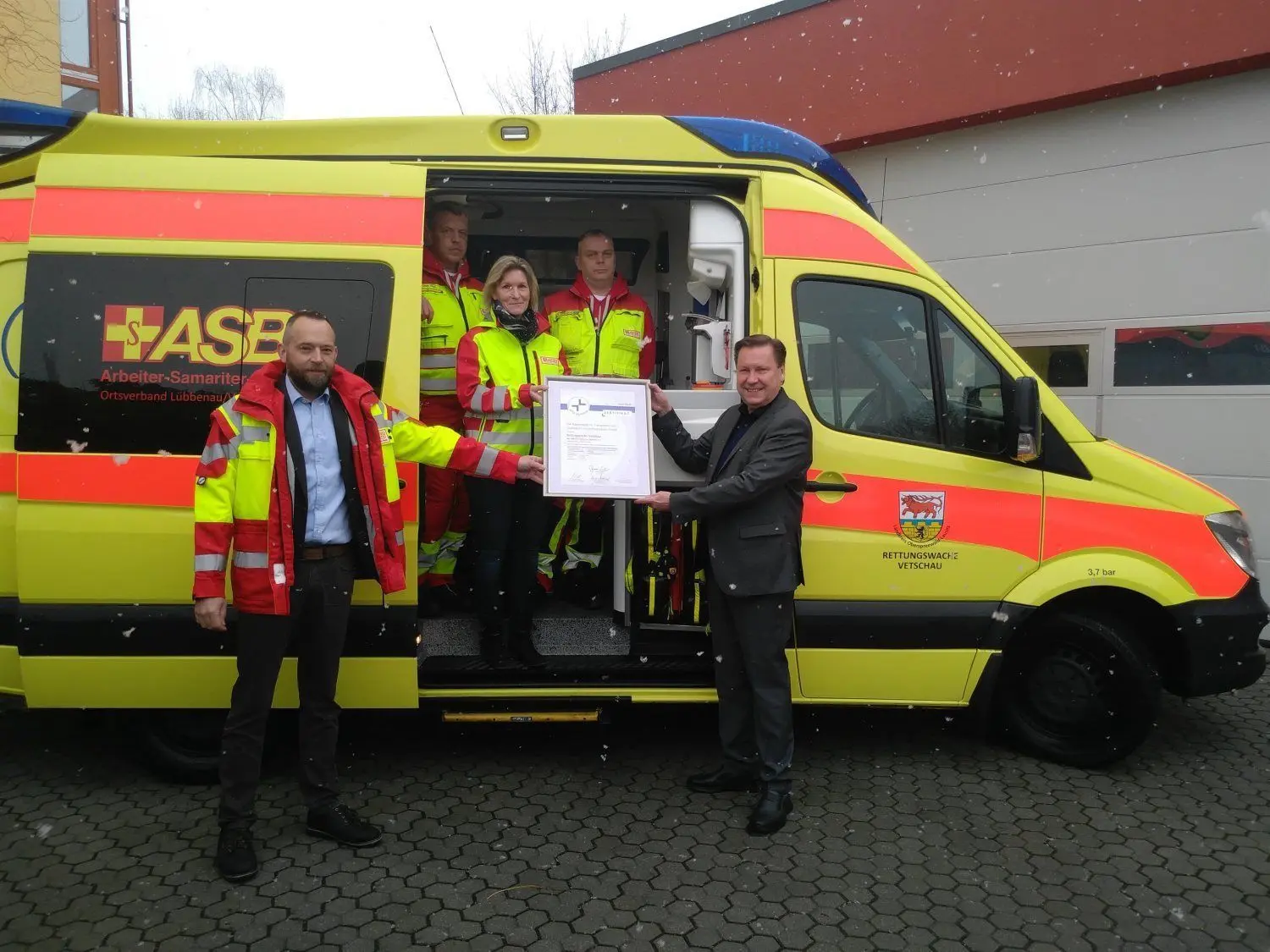 Ronald Neubauer (r.), Geschäftsführer von der KTQ GmbH, überreicht ASB-Geschäftsführer Alexander Kohl (l.) das Qualitätssiegel für die ASB-Rettungswache in Vetschau. Im Rettungswagen freut sich die Leiterin der Rettungswache, Simone Tartz, im Beisein von zwei Notfall-Sanitätern über die Auszeichnung.