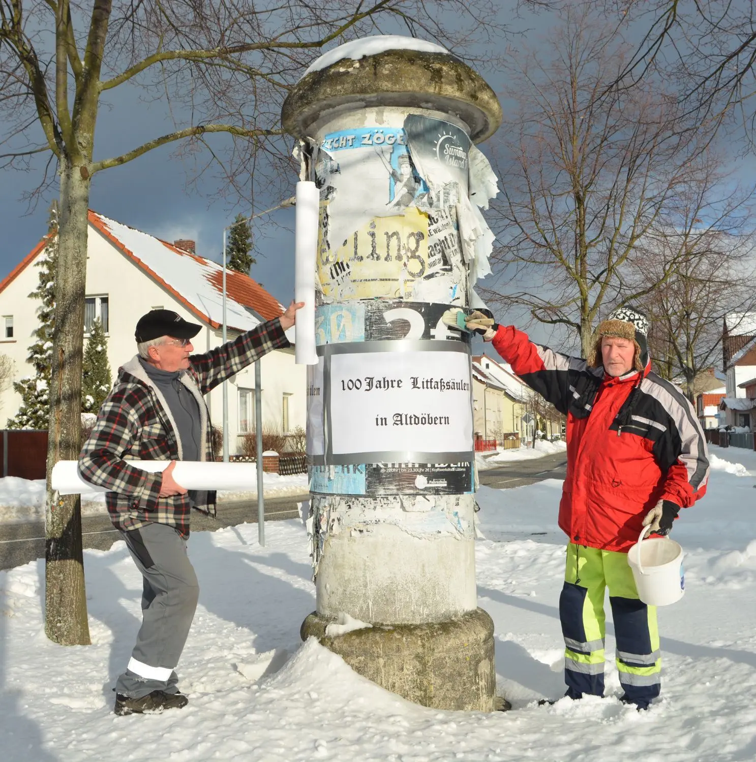 An den beiden verbliebenen Altdöberner Litfaßsäulen haben Klaus-Dieter Schmidt (l.) und Uwe Toppe (r.) Glückwunschbanderolen zum 100. Geburtstag angebracht.