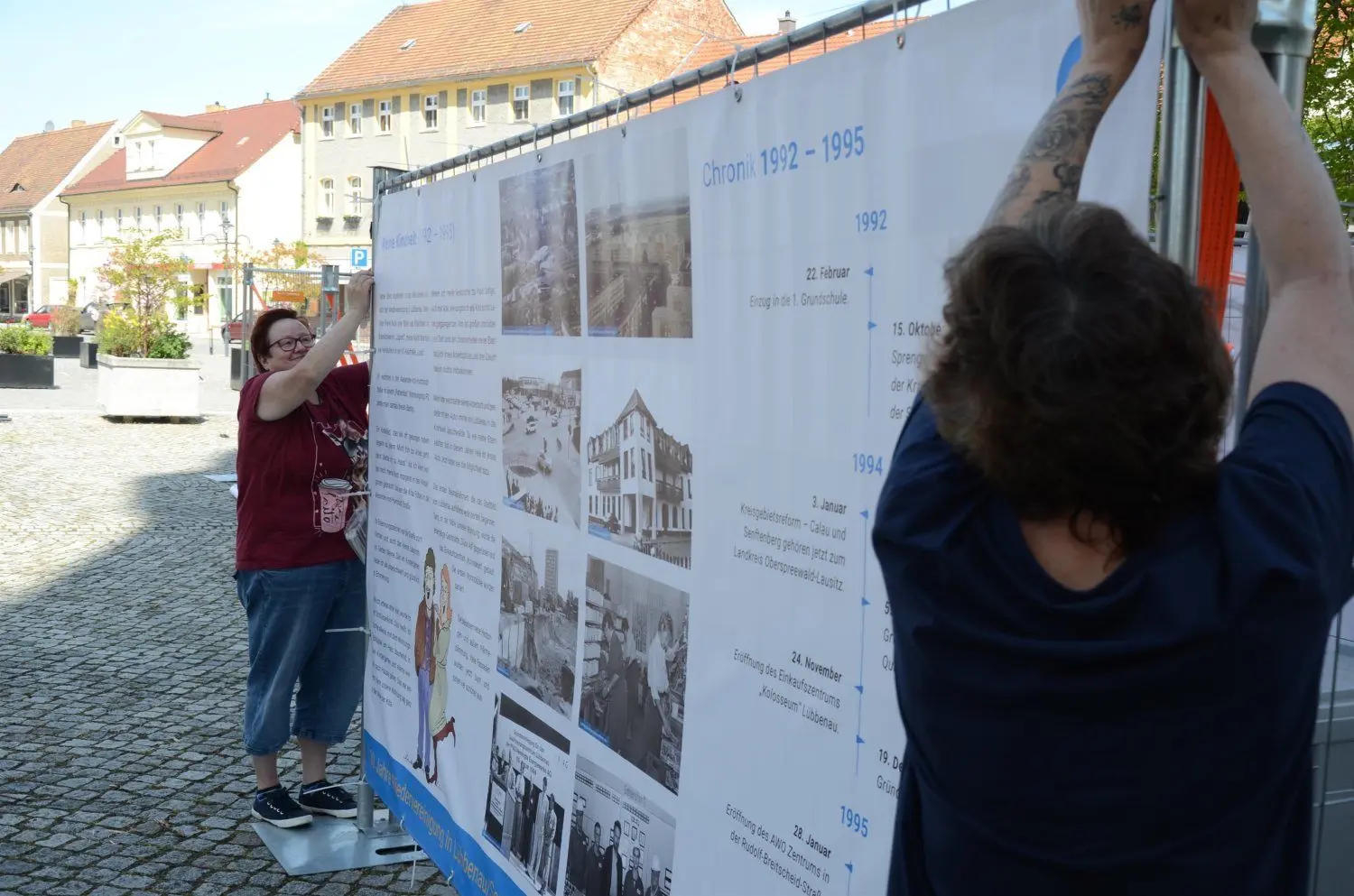 Gudrun Wilhelm und Cordula Milicic befestigen eins von insgesamt neun Erzählbannern auf dem Lübbenauer Kirchplatz.