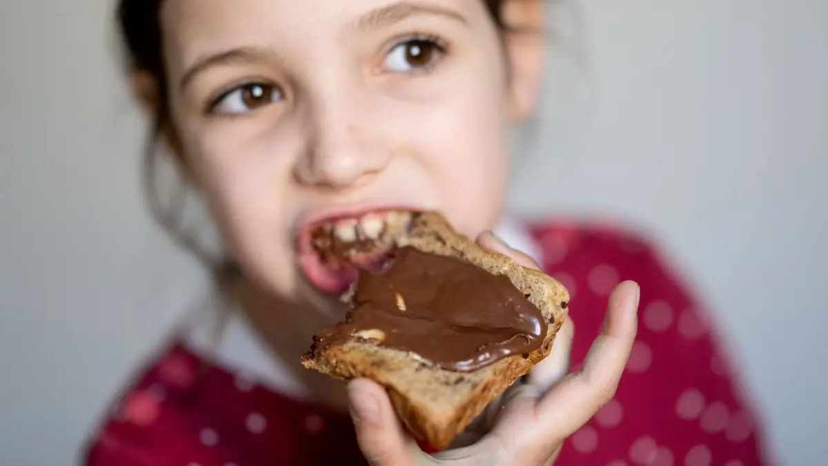 Nutella wird 60: ILLUSTRATION - 10.04.2024, Sachsen, Leipzig: Ein Mädchen isst ein Nutella-Brot. Der Brotaufstrich wird 60. Im italienischen Alba lief am 20. April 1964 das erste Glas vom Band. Erfunden wurde die Nuss-Nougat-Crème von Pietro Ferrero. Heute ist sie fast überall auf der Welt ein Erfolg. (zu dpa: «Die, der, das, mit oder ohne? Nutella wird 60») Foto: Hendrik Schmidt/dpa - ACHTUNG: Nur zur redaktionellen Verwendung im Zusammenhang mit der aktuellen Berichterstattung und nur mit vollständiger Nennung des vorstehenden Credits +++ dpa-Bildfunk +++