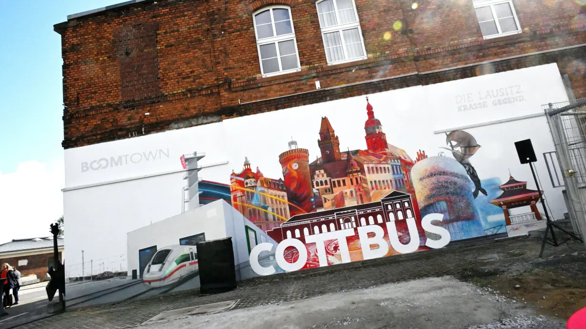 Wandbild Bunter Bahnhof: Graffiti-Kunst am Bunten Bahnhof
Der Cottbuser Künstler Dirk Hiekel hat die Fassade des Bunten Bahnhofes verschönert und ein 13 Meter langes großes Wandbild mit der Cottbuser Stadtsilhouette geschaffen. Dirk Hiekel, auch bekannt unter dem Namen „Koenichson“ hat im Auftrag der Deutschen Bahn (DB) auf einer Fläche von rund 60 Quadratmetern die Cottbuser Skyline neu in Szene gesetzt und um das in direkter Nachbarschaft gelegene ICE-Werk erweitert. Rund drei Wochen hat der Cottbuser Graffiti-Künstler an dem großformatigen Werk gearbeitet.
Neben dem Spremberger Turm, der Oberkirche St. Nikolai und dem Amtsgericht reihen sich auf dem großflächigen Wandbild unter anderem das Wappentier der Stadt Cottbus, der Krebs, und die Kunsthalle Lausitz sowie der Aussichtsturm Merzdorf am Cottbuser Ostsee ein. Die Schriftzüge der Image-Kampagnen der Stadt Cottbus „Boomtown Cottbus“ sowie der Wirtschaftsregion Lausitz „Die Lausitz. Krasse Gegend“ runden das Kunstwerk ab. Während der feierlichen Übergabe des Wandbildes betonte Ministerin Kathrin Schneider, dass das Werk die enge Zusammenarbeit zwischen der Staatskanzlei des Landes Brandenburg, der Stadt Cottbus und der Deutschen Bahn für den Strukturwandel in der Lausitz symbolisiere.