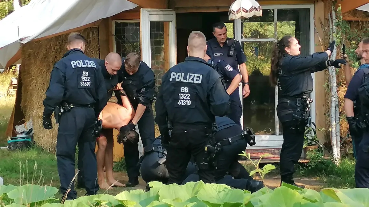 Zwei Bewohner haben sich geweigert, die selbst gebaute Unterkunft zu verlassen. Daraufhin wird eine Frau am Boden fixiert, der Eigentümer in unbequemer Haltung zur Seite geführt.