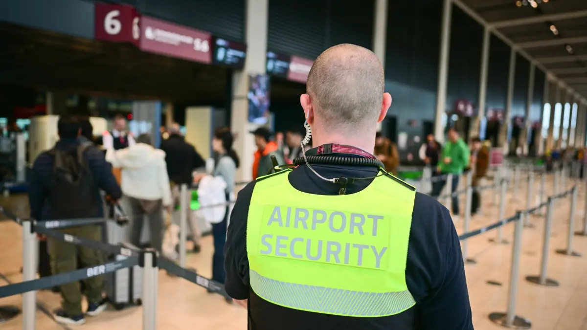 Flughafen BER Reiseverkehr vor Ostern: 09.04.2025, Brandenburg, Schönefeld: Ein Sicherheitsmitarbeiter mit der Aufschrift «Airport Security» auf seiner Weste steht während eines Pressetermins am Flughafen Berlin Brandenburg im Terminal 1. Im südlichen Teil des Terminal 1 sind auf den Sicherheitspuren nun sogenannten CT-Scannern im Einsatz. Diese sollen die Kontrollen beschleunigen. Foto: Sebastian Christoph Gollnow/dpa +++ dpa-Bildfunk +++