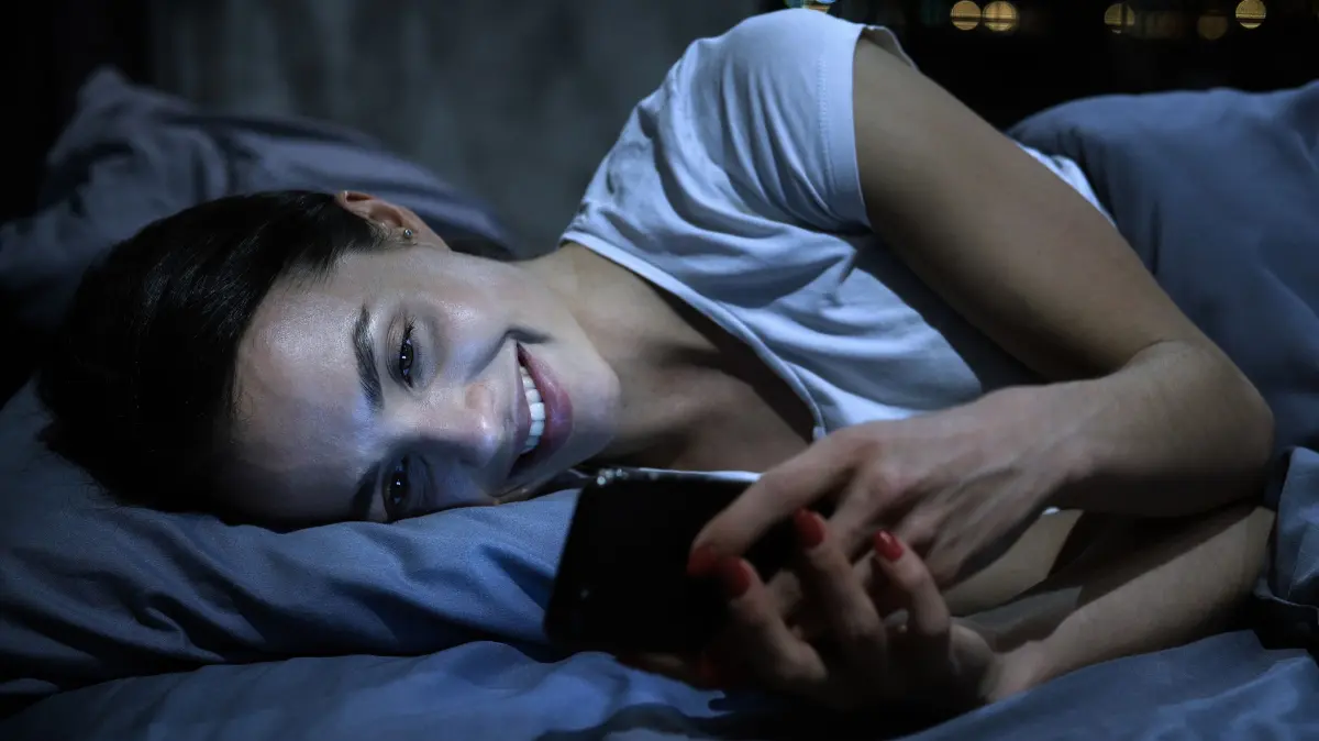 Young beautiful smiling woman using mobile phone in bed at dark bedroom at night