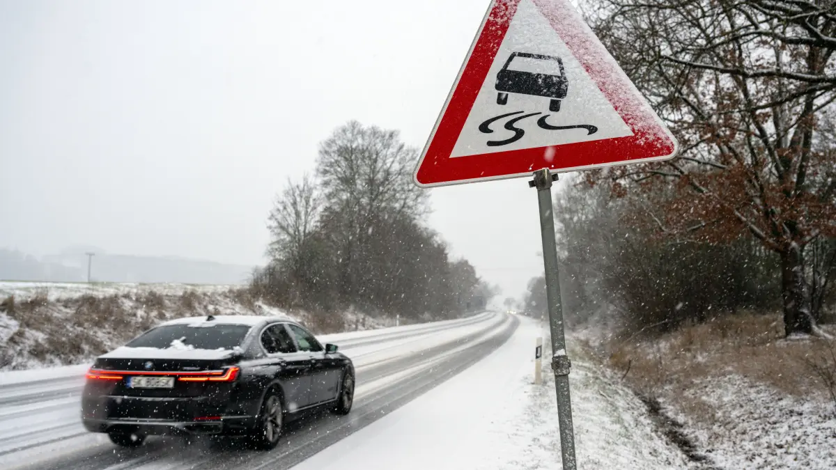 Ein Auto fährt auf einer mit Schnee bedeckten Straße, an der ein Schild vor Rutschgefahr warnt.