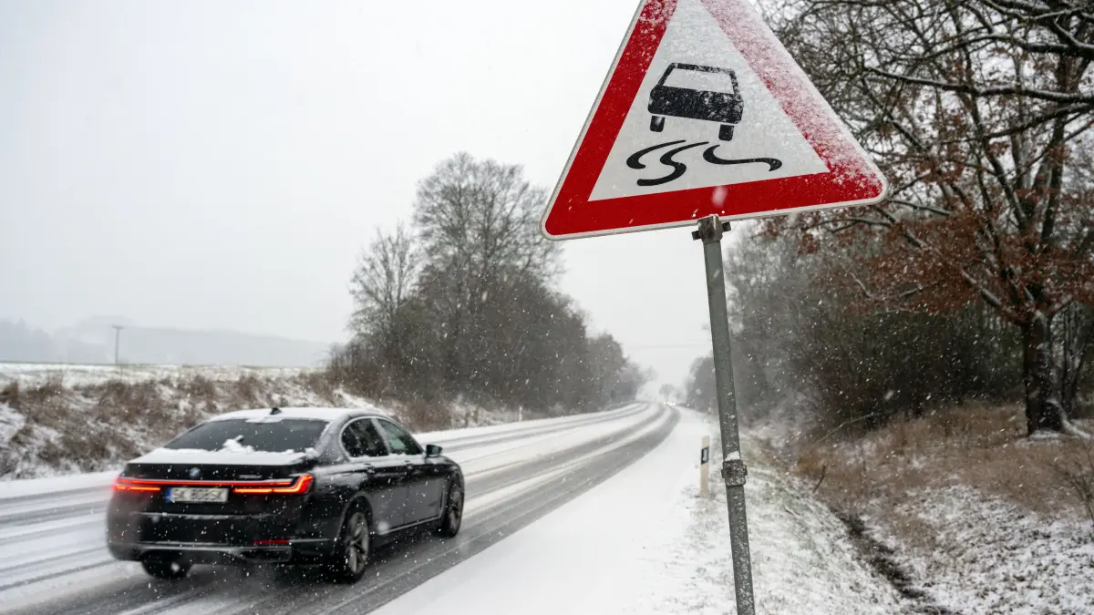 Ein Auto fährt auf einer mit Schnee bedeckten Straße, an der ein Schild vor Rutschgefahr warnt.