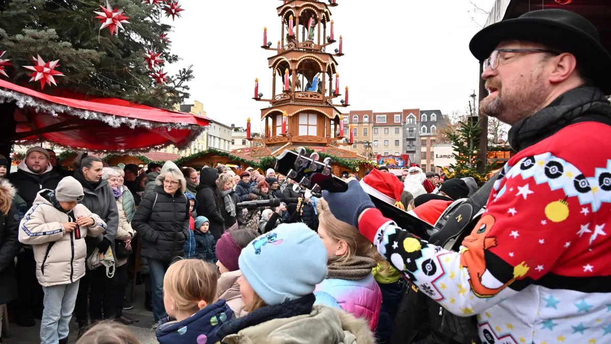 Weihnachtsmarkt 2025 Stollenanschnitt: Torsten Karow sang mit den Kindern vom Sachsendorfer Kinderchor sowie dem Chor der Bauhausschule Weihnachtslieder.
Stollenanschnitt zur Eröffnung des Weihnachtsmarktes
Am Montag ist auf dem Altmarkt mit dem traditionellen Stollenanschnitt der diesjährige Weihnachtsmarkt der 1000 Sterne eröffnet worden. Gemeinsam mit dem Weihnachtsmann und Vertretern der Cottbuser Rathausspitze ist der handwerklich perfekt hergestellte sechs Meter lange, 35 bis 40 Kilogramm schwere und mit 20 Kilogramm Butter gebackene Butter-Rosinenstollen zur Eröffnung des Weihnachtsmarktes der 1.000 Sterne angeschnitten worden. In diesem Jahr stellten die Weihnachtsstollen die Bäckerei Dreißig aus Guben, Die Bäckerei Bubner aus Doberlug – Kirchhain, die Bäckerei Arlt aus Cottbus, die Bäckerei Wahn aus Vetschau sowie die Bäckerei Hanuschka aus Gallinchen die meisterlich hergestellten einzelnen Stollen zur Verfügung. Die Innung der Lausitzer und Spreewälder Bäcker- und Konditoren an der hält schon viele Jahre an dieser Tradition zur Eröffnung des Cottbuser Weihnachtsmarktes fest und freute sich über Spenden für jedes Stück Weihnachtstollen. Die Spende pro abgegebenes Stück geht in diesem Jahr wieder an das Kinderhaus „Pusteblume“ in Burg. Die Innung wird den tatsächlichen Spendenerlös aufrunden, bevor er an das Kinderhaus übergeben wird.