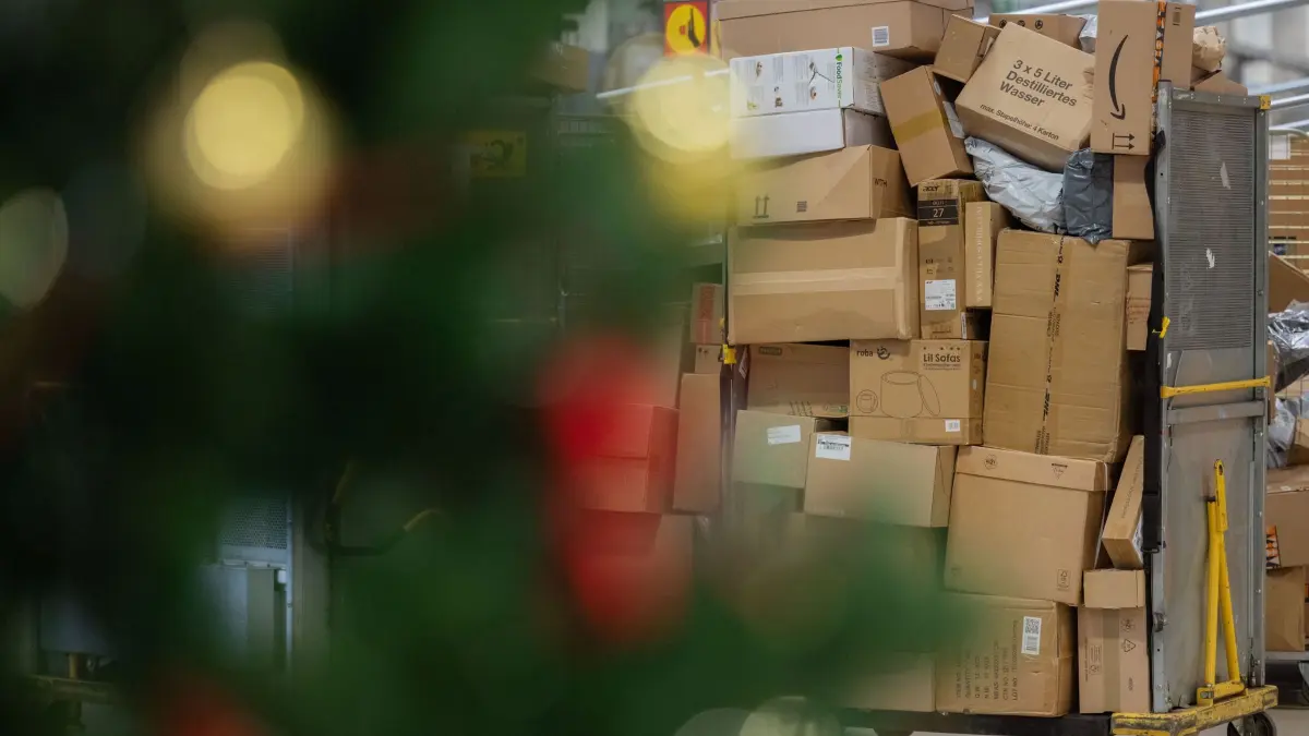 Paketbeförderung in der Vorweihnachtszeit: PRODUKTION - 04.12.2025, Nordrhein-Westfalen, Köln: Pakete sind in einer Zustellbasis von Deutsche Post DHL hinter einem Weihnachtsbaum zu sehen. Aktuell fallen durch den Black Friday (Black Week) und Weihnachten mehr Pakete an. Im diesjährigen Weihnachtsgeschäft rechnet die Deutsche Post DHL wieder mit Paketmassen. Foto: Rolf Vennenbernd/dpa +++ dpa-Bildfunk +++