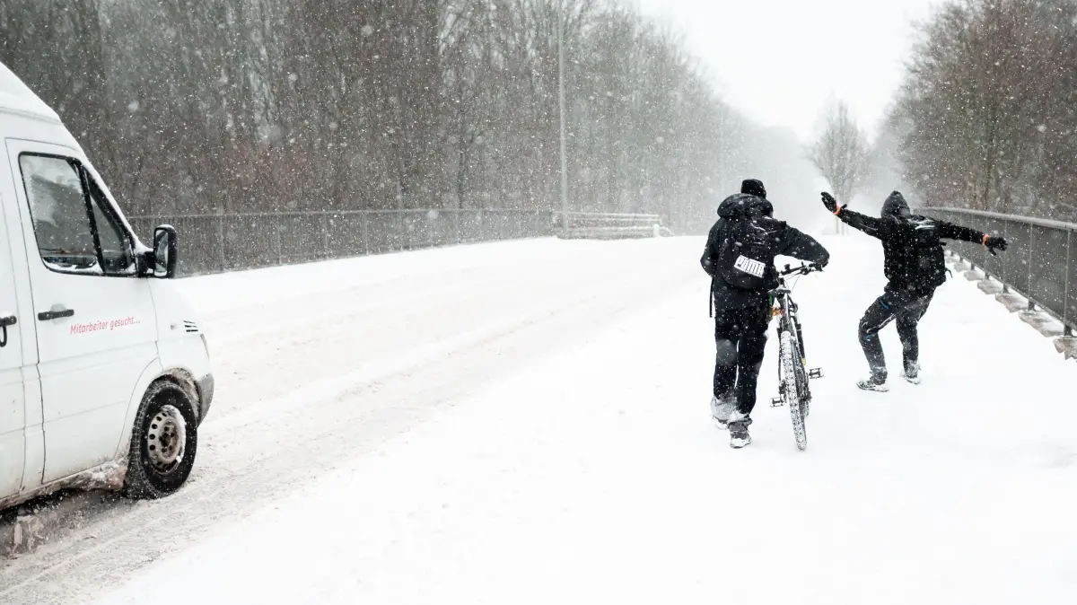 Ein Fahrradfahrer schiebt sein Rad an einer Straße entlang während ein Mann daneben im Schnee rutscht. Die starken Schneefälle in Nordrhein-Westfalen führen zu starken Einschränkungen im Verkehr. +++ dpa-Bildfunk +++