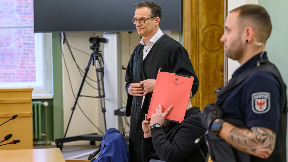 Nach Tod eines Polizisten in Lauchhammer - Prozess beginnt: ARCHIV - 14.01.2026, Brandenburg, Cottbus: In Handschellen und Aktenordner vor dem Gesicht sitzt der Angeklagte im Mordprozess um einen getöteten Polizisten aus Dresden neben seinem Verteidiger Christoph Rühlmann zum Prozessbeginn in einem Verhandlungssaal des Cottbuser Landgerichts. Bei einem Einsatz gegen flüchtende Autodiebe wird ein Polizist aus Dresden in Südbrandenburg von einem Wagen erfasst und getötet. Rund ein Jahr später verhandelt das Landgericht Cottbus wegen des Vorwurfs des Mordes gegen den Fahrer eines gestohlenen Wagens. (zu dpa: «Tod von Polizisten: Angeklagter fuhr wie ein «Rallyefahrer»») Foto: Patrick Pleul/dpa +++ dpa-Bildfunk +++