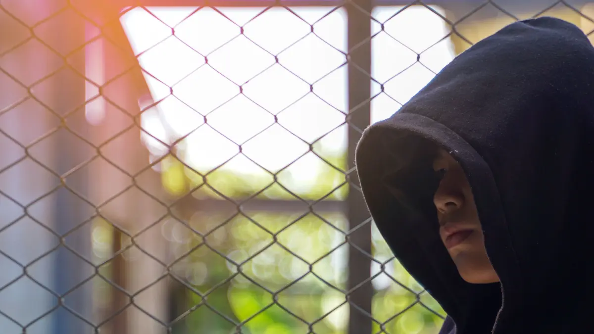 NPG-erweiterte Lizenz / Mantel, Ein geheimnisvoller Teenager in einem schwarzen Kapuzenpullover steht vor einem Maschendrahtzaun. Das Thema thematisiert Jugendkriminalität und Anonymität in der Stadt. 1767746014 Download am 17.3.26 für SWP, SWUM. Foto: © Aoy_Charin/adobe.stock.com