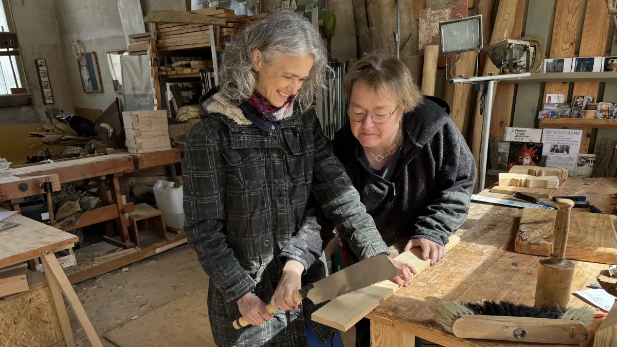 Im Atelier von Alma Fichtner auf dem Teluxgelände in Weißwasser. Ines Forgel-Sedlmeir (l.) und Alma Fichtner (r.) an der Werkbank mit einer Holzsäge. Alma zeigt ihr, wie sie das Holzbrett richtig zersägen kann.