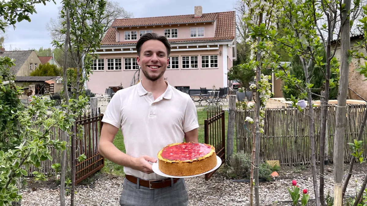 Kendrick Kling posiert am 24.04.2026 im Kräutergarten des Cafés Lavendel, Glück und Landgenuss in Schwarzheide. Er hält "Omas Käsekuchen mit Erdbeeren" in die Kamera.