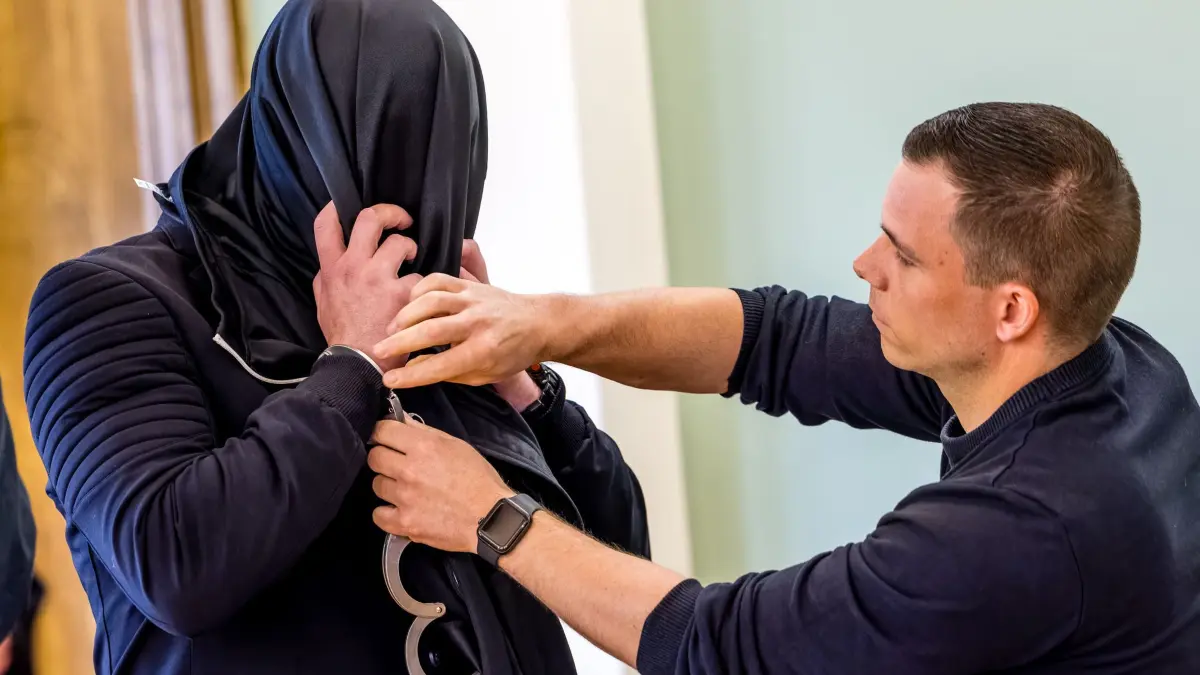 Prozess am Landgericht Cottbus wegen Mordes an einem Polizisten: 06.05.2026, Brandenburg, Cottbus: Dem Angeklagten (l) werden von einem Justizangestellten die Handfesseln abgenommen. Dem Angeklagten wird Mord an einem 32 jährigen Polizisten vorgeworfen. Bei einem Einsatz gegen mutmaßliche Autodiebe vor einem Jahr in Lauchhammer war dieser von einem Fahrzeug erfasst worden und noch am Einsatzort gestorben. Foto: Frank Hammerschmidt/dpa +++ dpa-Bildfunk +++