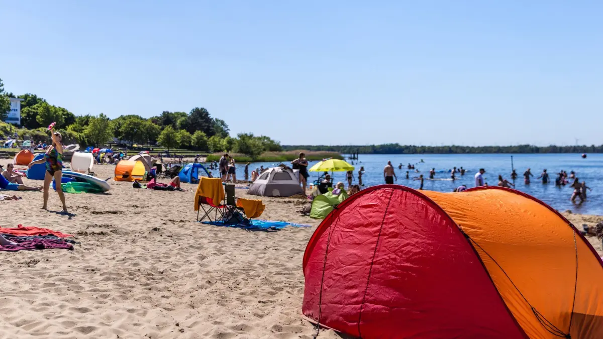 Sommerliches Wetter am Pfingstmontag: 25.05.2026, Brandenburg, Senftenberg: Sonnenschirme und Zelte stehen am Großkoschener Seestrand und Menschen baden im Senftenberger See. Die Temperaturen in Südbrandenburg werden heute die 30-Grad-Marke nicht erreichen, die Sonne scheint aber ganztägig und es bleibt trocken. Foto: Frank Hammerschmidt/dpa - ACHTUNG: Verwendung nur im vollen Format +++ dpa-Bildfunk +++