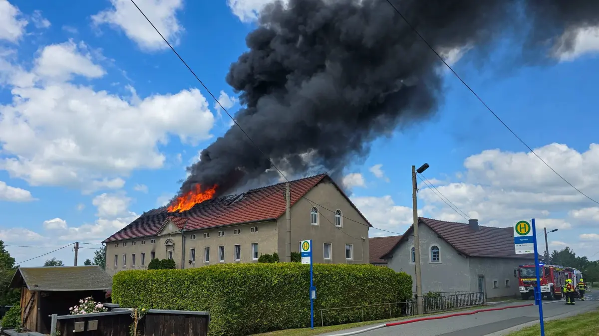 Datum: 30.05.2026 / Ort: Eiserode
Seit Samstagmittag sorgt ein Brand in einem Mehrfamilienhaus in Eiserode bei Loebau fuer einen Grosseinsatz der Rettungskraefte. Nach ersten Informationen war das Feuer zunaechst im Gebaeude ausgebrochen, ehe sich die Flammen rasch ausbreiteten und schliesslich durch das Dach schlugen.
Eine dichte schwarze Rauchwolke zog ueber den Ort hinweg und war bereits aus groesserer Entfernung zu sehen. Zahlreiche Einsatzkraefte der Feuerwehr sind vor Ort, um den Brand unter Kontrolle zu bringen und eine weitere Ausbreitung der Flammen zu verhindern.
Zur Brandursache sowie zur Hoehe des entstandenen Sachschadens liegen derzeit noch keine gesicherten Erkenntnisse vor. Auch Angaben zu moeglichen Verletzten standen zunaechst aus. Die Loescharbeiten dauern an.