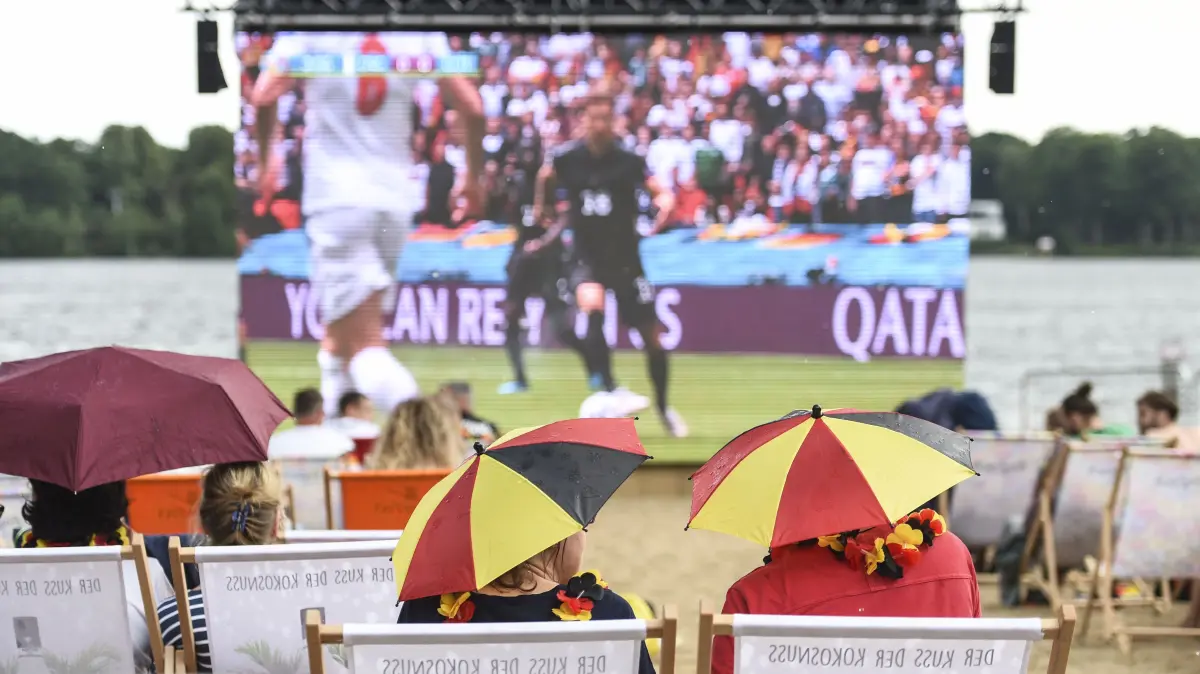 Besucherinnen und Besucher sitzen mit Schirmen in den Farben der deutschen Flagge als Hüte am Strandbad Wendenschloss beim Public Viewing während des Spiels der deutschen Mannschaft gegen England im Achtelfinale. +++ dpa-Bildfunk +++