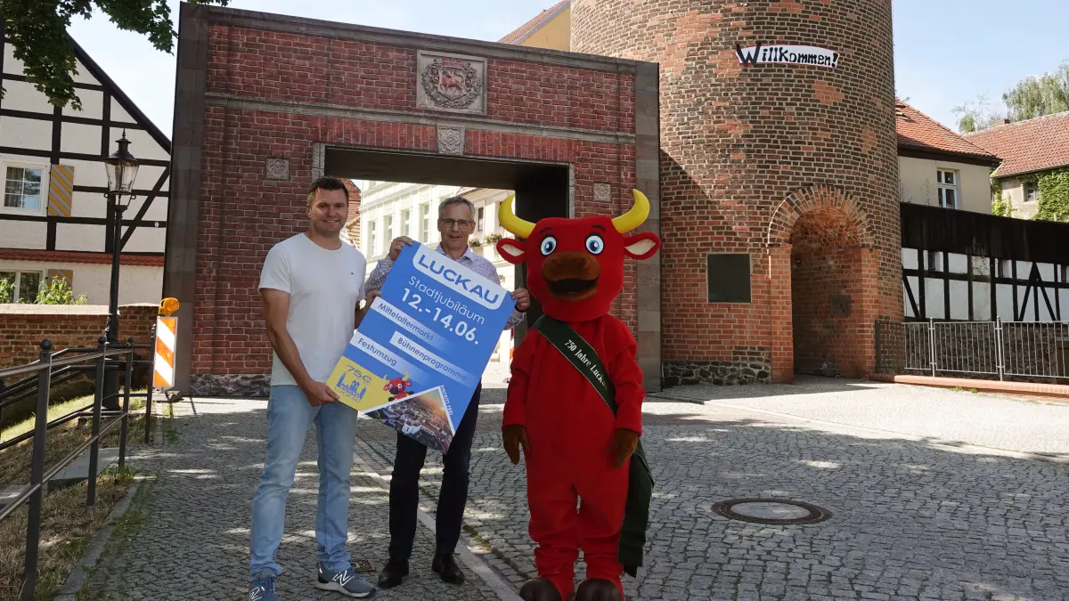 Luckau lädt zum großen Festwochenende anlässlich der 750-Jahr-Feier von Luckau am 12., 13. und 14. Juni 2026. Tino Wolff (Landesgartenschau Luckau 2000 gGmbH), Gerald Lehmann (Bürgermeister Luckau) & das Maskottchen Lucki stehen vor einem Wahrzeichen der Stadt, dem Roten Turm, und der Rekonstruktion des ehemaligen Stadttores von Luckau.