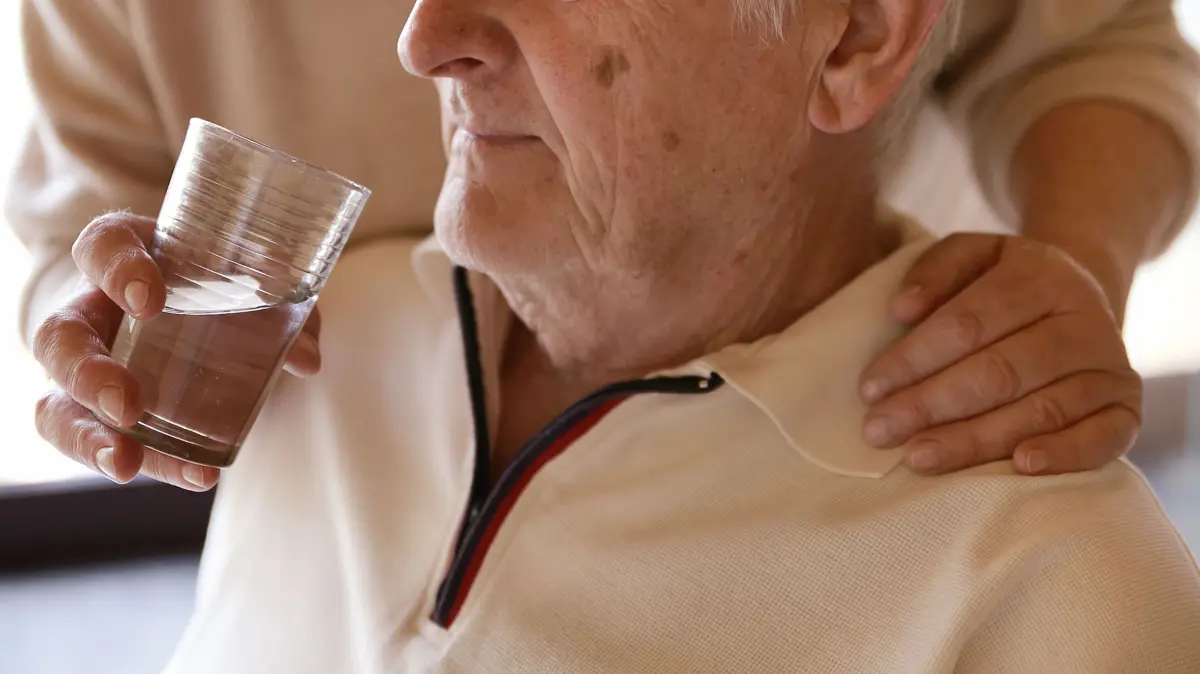 Eine Frau hilft einem Mann beim Trinken: ILLUSTRATION - Sich als Pflegeperson eintragen zu lassen, bringt nur Vorteile - eine bessere Absicherung im Alter und bei Unfällen etwa. (zu dpa: «Als Pflegeperson eintragen lassen - was bringt das?») Foto: Mascha Brichta/dpa-tmn - Honorarfrei nur für Bezieher des dpa-Themendienstes +++ dpa-Themendienst +++