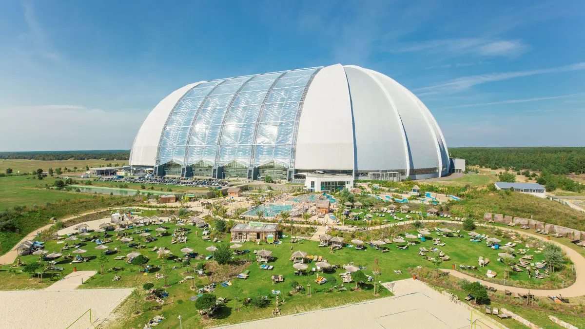 Blick auf die Kuppel von Tropical Islands. Im Vordergrund: Der Außenbereich "Amazonia" mit Poollandschaft, zahlreichen Wasserattraktionen, sowie ausgedehnten Liege- und Sportflächen.