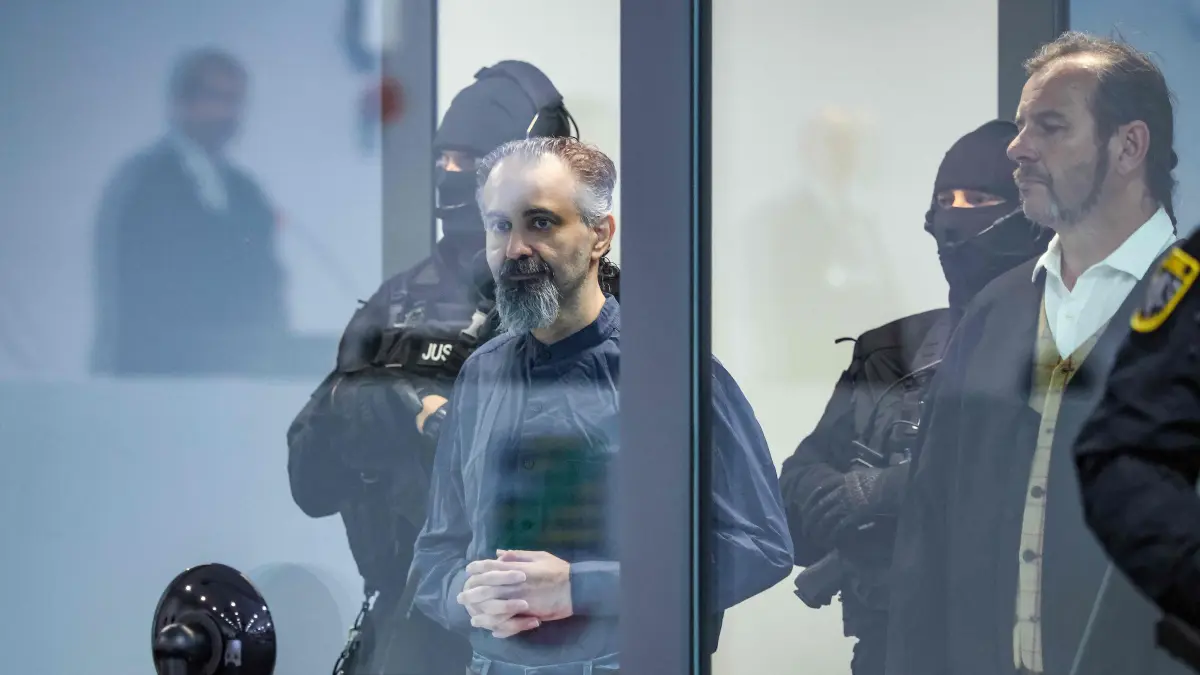GERMANY-COURT-CRIME-ATTACK: Saudi Arabian defendant Taleb Jawad al-Abdulmohsen (C) stands next to a judicial security officer as presiding judge Dirk Sternberg (L) is reflected in a bulletproof glass box in the temporary courtroom in Magdeburg, eastern Germany, before al-Abdulmohsen's trial for driving an SUV through a crowd at a Christmas market. The court sentenced to life in jail the Saudi psychiatrist who drove an SUV through a crowded Christmas market in 2024, killing six people and injuring more than 300. (Photo by Ronny HARTMANN / AFP)