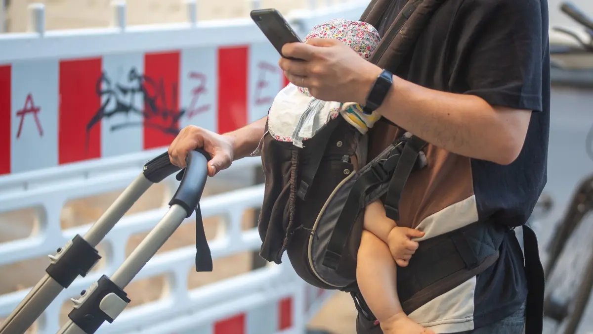 Elterngeld: SYMBOLBILD - 09.09.2023, Berlin: Ein Mann schiebt einen Kinderwagen und trägt gleichzeitig ein Baby in einer Tragehilfe. Dabei schaut er auf sein Smartphone. (zu dpa: «Welche Einschnitte beim Elterngeld? Entscheidung Anfang Juli») Foto: Fernando Gutierrez-Juarez/dpa +++ dpa-Bildfunk +++