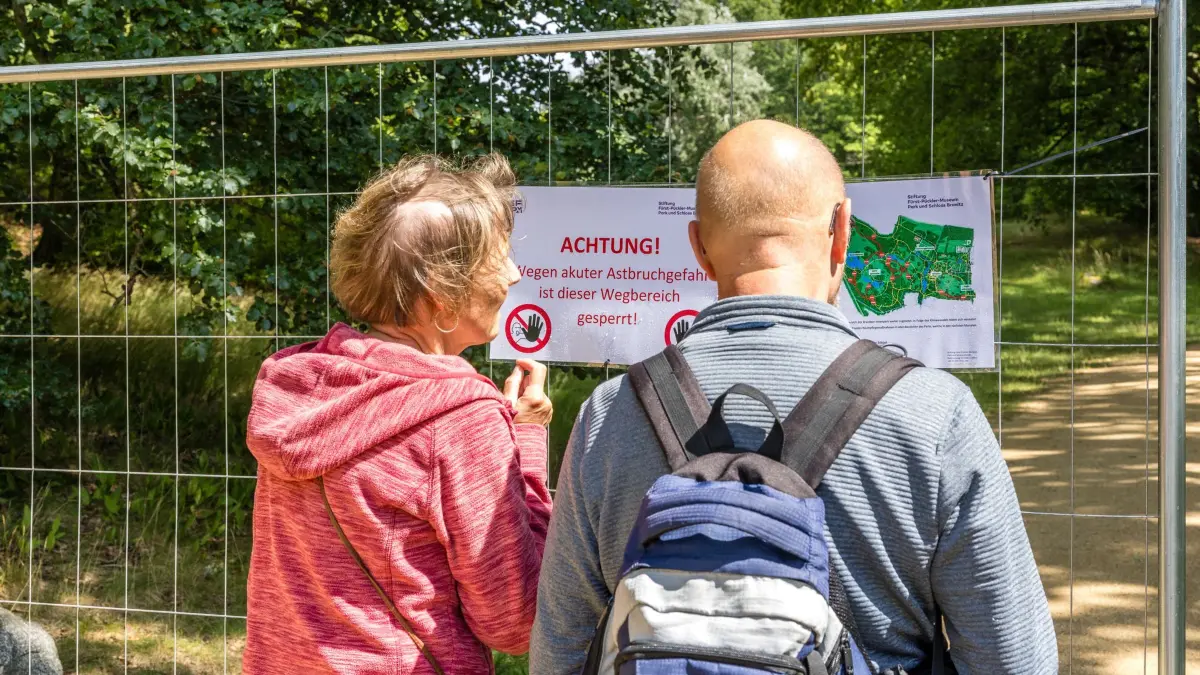 Sperrungen im Branitzer Park: 20.07.2026, Brandenburg, Branitz: Eine Frau und ein Mann lesen Schilder, die an einem Zaun angebracht sind und auf gesperrte Wege hinweisen. Aufgrund akuter Gefahr von Astabbrüchen sind seit heute einige Wege im Branitzer Park gesperrt. Für Besucher stehen aber weiterhin viele Wege zwischen den Branitzer Sehenswürdigkeiten offen. Foto: Frank Hammerschmidt/dpa +++ dpa-Bildfunk +++