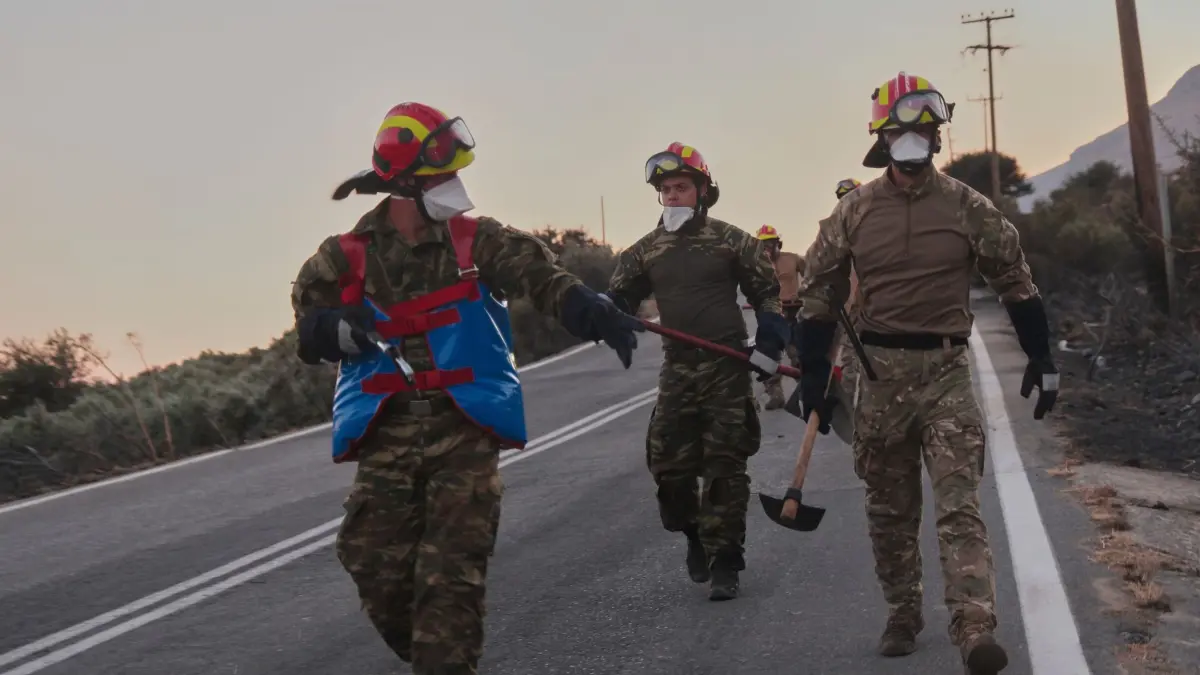 Waldbrände in Griechenland - Kreta: 29.07.2026, Griechenland, Kreta: Feuerwehrleute gehen während eines Waldbrands in der Region Rethymno auf der südgriechischen Insel Kreta mit Werkzeugen eine Straße entlang. Foto: Giannis Angelakis/AP/dpa +++ dpa-Bildfunk +++