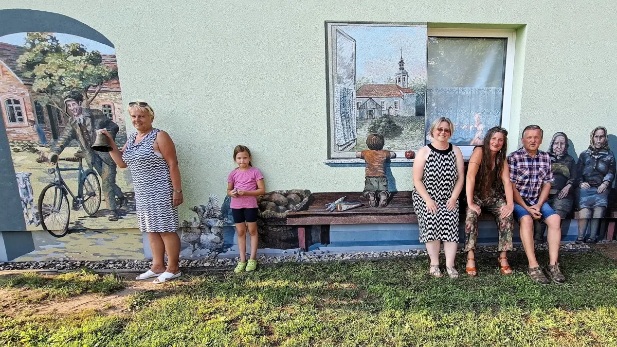 Das Bild erzählt Dorfgeschichten entlang des Lausitzer Landweges: Auf dem Wandbild der Klingelmann mit Gabi Rostock samt Glocke davor. Auf der gemalten Bank haben sich „sitzend“ Ortsvorsteherin Silvana Lehmann, die Künstlerin Stephanie Mai und Wilfried Lange, Vorsitzender des Traditionsvereins, dazugesellt.