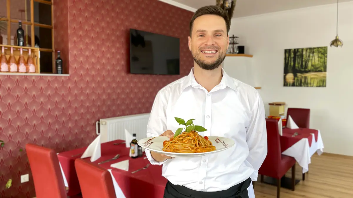 Vedad Selami, von Freunden Vedi genannt, steht am 6. August 2026 mit einem Teller Spaghetti mit Garnelen und Tomatensoße in der Hand in seinem Restaurant Trattoria La Piazza in Golßen.