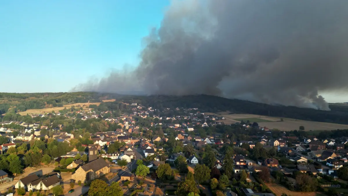 Waldbrand im Hürtgenwald: 14.08.2026, Nordrhein-Westfalen, Hürtgenwald: Dichter Rauch eines Waldbrands im Hürtgenwald zieht über den Ortsteil Gey (Aufnahme mit Drohne). Am frühen Morgen hat die Evakuierung des Ortsteils Gey begonnen, in dem gut 1.800 Menschen leben. Zuletzt war gemeldet worden, dass die Flammen rund 400 Meter von dem Ort entfernt seien. Foto: Federico Gambarini/dpa +++ dpa-Bildfunk +++