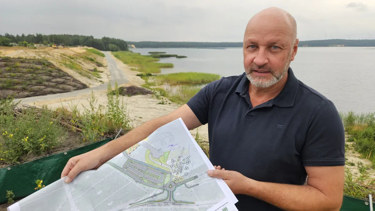 Projektmanager Steffen Mühl zeigt auf dem Plan, wie das Areal am Scheibe-See künftig aussehen soll. Im Hintergrund sind die entstehenden Terrassen und die Zufahrt zur späteren Mole zu sehen.