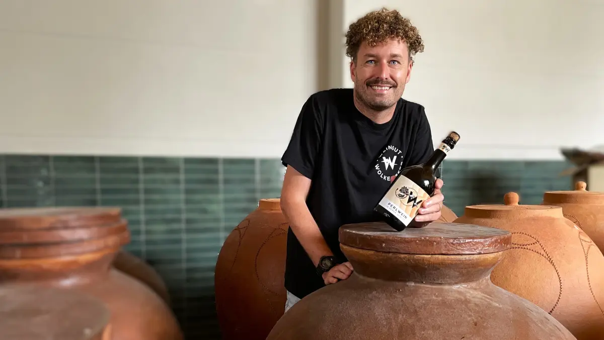 Philipp Henke, Vertriebsleiter des Weingutes Wolkenberg, hält am 17.08.2026 eine Flasche Perlwein in die Kamera. Er steht im Lager zwischen den neuen Dergas.