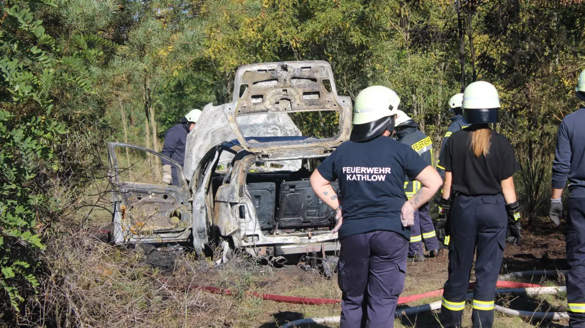 Brand bei Cottbus: In einem Waldstück bei Kathlow in Neuhausen/Spree hat ein Pkw gebrannt. Feuerwehrleute stehen am gelöschten Autowrack.